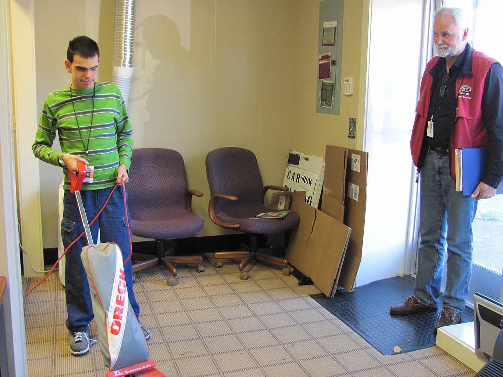 Christopher Merritt vacuums floors at Camas Technology Associates. Customers walking into the business are greeted with a big smile and handshake, courtesy of the CHS intern. Looking on is Henry Midles, Life Skills teacher.