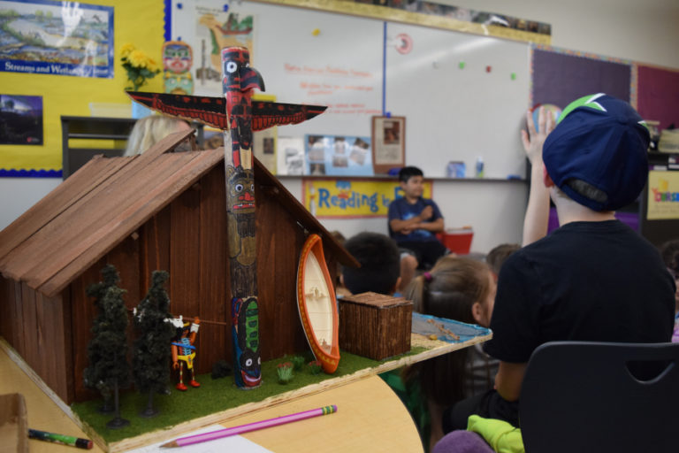 Third grade student Mason Taylor sits alongside his plank house while classmate Dereck Rodriguez Hernandez presents his project to the class.