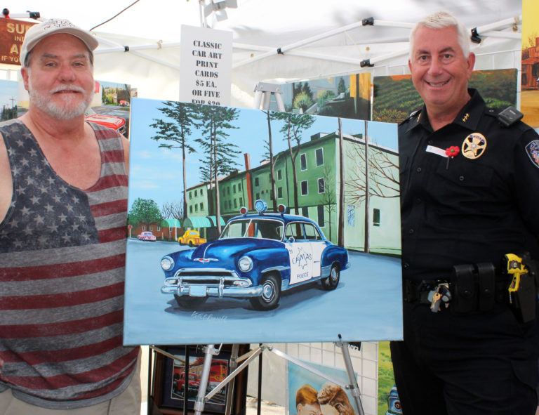Camas artist Keith E. Russell (left) and Camas Police Chief Mitch Lackey (right) stand next to Russell's painting of an early 1950s Camas Police car in front of the old Camas Hotel. Russell plans to donate the painting to the Camas Police Department. Lackey said he's been looking forward to seeing the finished work and was pleasantly surprised when he viewed it for the first time on Friday, July 27, during the first day of the 2018 Camas Days celebration. To find more of Russell's work, visit Facebook.com/ReflectionpaintingsbyKeith, or email KRussell@Maxcessintl.com. The Camas artist is open to doing commissioned pieces and specializes in paintings of classic vehicles.