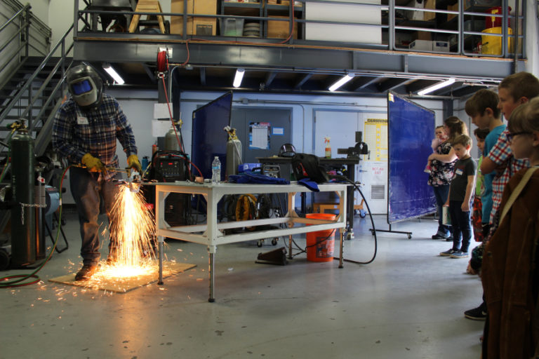 SIGMADESIGN welder Ryan Zahniser (left) shows several elementary school aged children of SIGMADESIGN employees how his welding torch will cut through steel at the product design and engineering firm&#039;s Camas headquarters off Northwest Pacific Rim Boulevard on April 25 while his wife, Michelle (second from left) and 2-year-old daughter, Madi, look on.