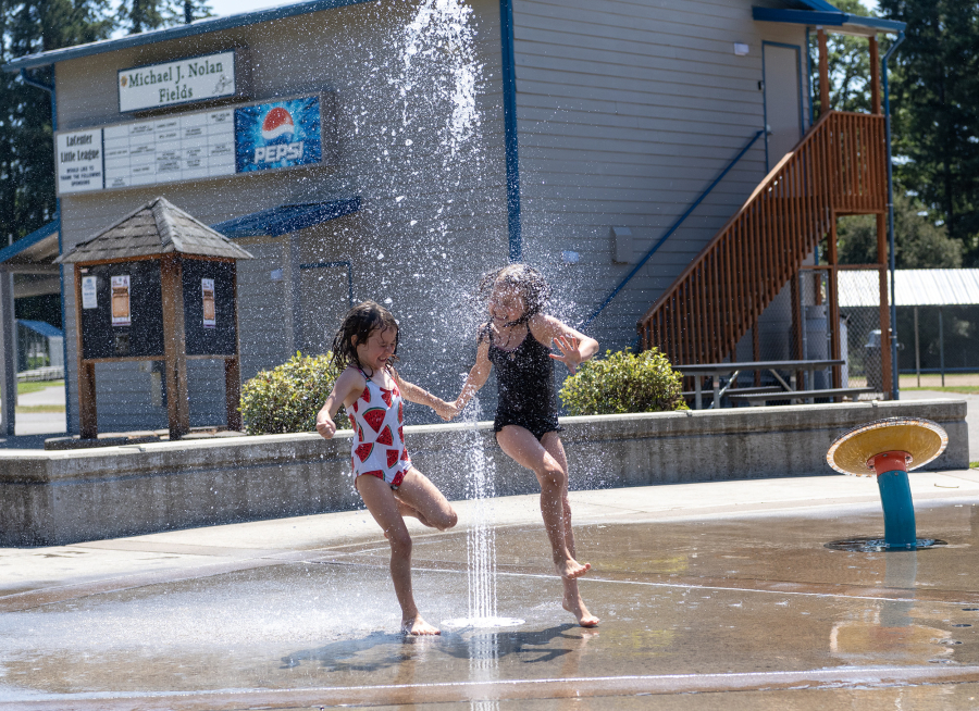 Emily, 8, right, and Alice Cash, 5, jump through a sprinkler Tuesday at the Holley Park splash pad in La Center. Public pools require lifeguards, heating, treatment, use fees and more to operate, whereas splash pads use less water and don’t require lifeguards. (Photos by Taylor Balkom/The Columbian)