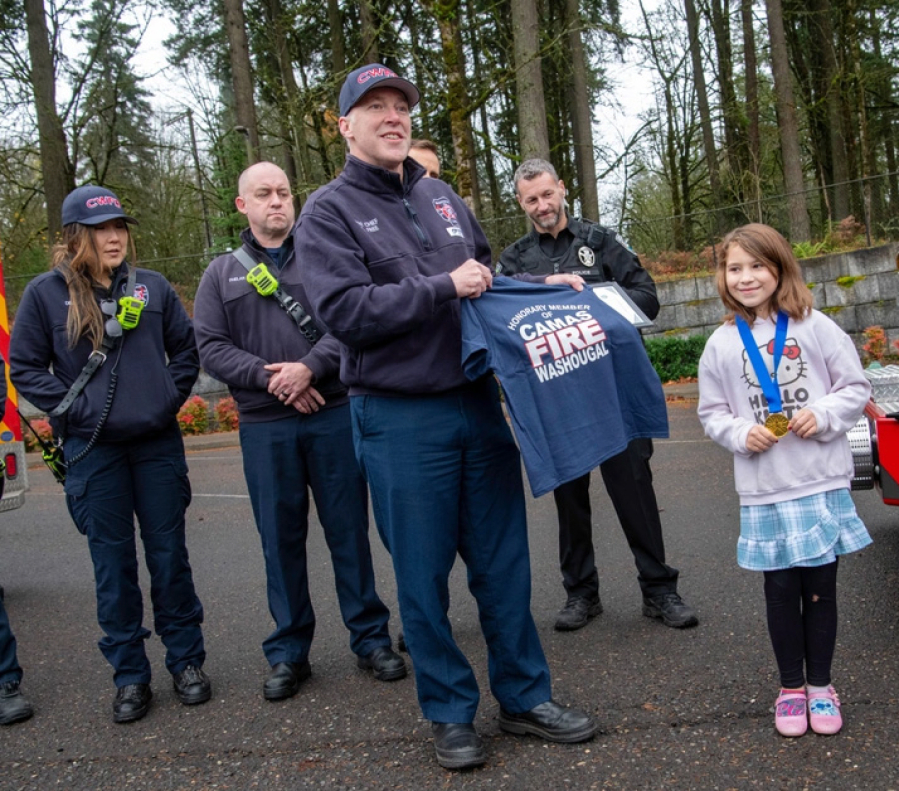 Camas-Washougal Fire Department Chief Cliff Free gives Shelbie McQueen, a third-grader at Woodburn Elementary School, an &ldquo;Honorary CWFD member&rdquo; T-shirt during an awards presentation held at the school Dec. 3. &ldquo;She wears that shirt all the time,&rdquo; said Shelbie&rsquo;s mother, Tiesha McQueen. &ldquo;She thinks it&rsquo;s really cool.&rdquo; (John Hughel/Oregon Military Department)