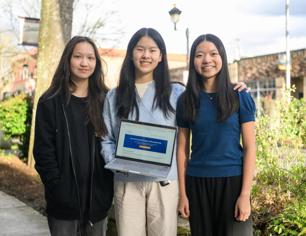 Camas High School sophomores Liya Zhao, from left, Chloe Luo and Alyssa Wong stand for a portrait with the home page of their cancer screening app pulled up Wednesday in downtown Camas. The trio were named the winners of the 2025 Congressional App Challenge for Washington&rsquo;s 3rd District. (Taylor Balkom/The Columbian)