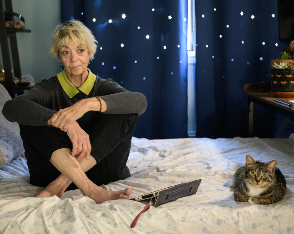 Home care aide Vicki Bickford sits on her bed for a portrait with her cat Binker on Tuesday at her home in Vancouver. Bickford and other advocates say WA Cares, a first-in-the-nation public long-term care benefit, fills a gap in long-term care not covered by Medicare or other forms of health insurance. The benefit also is available to pay family members to be caregivers. (Taylor Balkom/The Columbian)