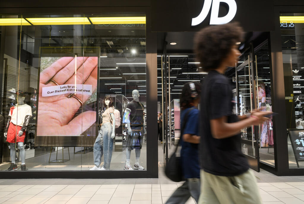 Shoppers walk in front of JD Sports on Aug. 25 at Vancouver Mall. At top, backpacks and other clothing items hang inside JD Sports. (Photos by Taylor Balkom/The Columbian)