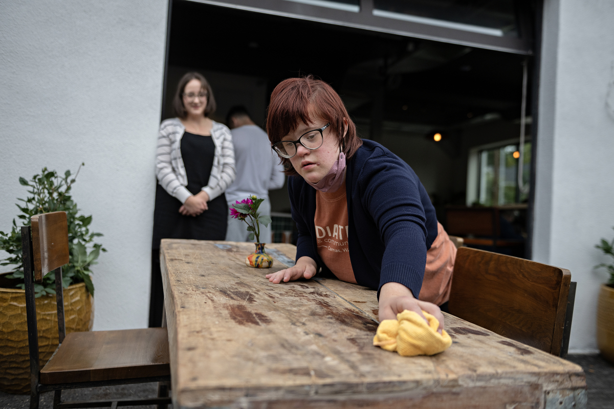 Job coach Jennifer Trout, left, looks on as Daria Eremenko of Washougal keeps the tables at Radiate Coffee and Community in top shape on the morning of Sept. 30. Eremenko said she enjoys the variety of tasks she can do and loves meeting customers. (Amanda Cowan/The Columbian)